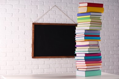 Photo of Stack of many colorful books on white wooden table and blackboard in classroom. Space for text