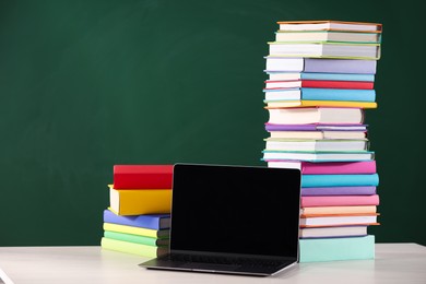 Photo of Stacks of many colorful books and laptop on white table near chalkboard in classroom. Space for text