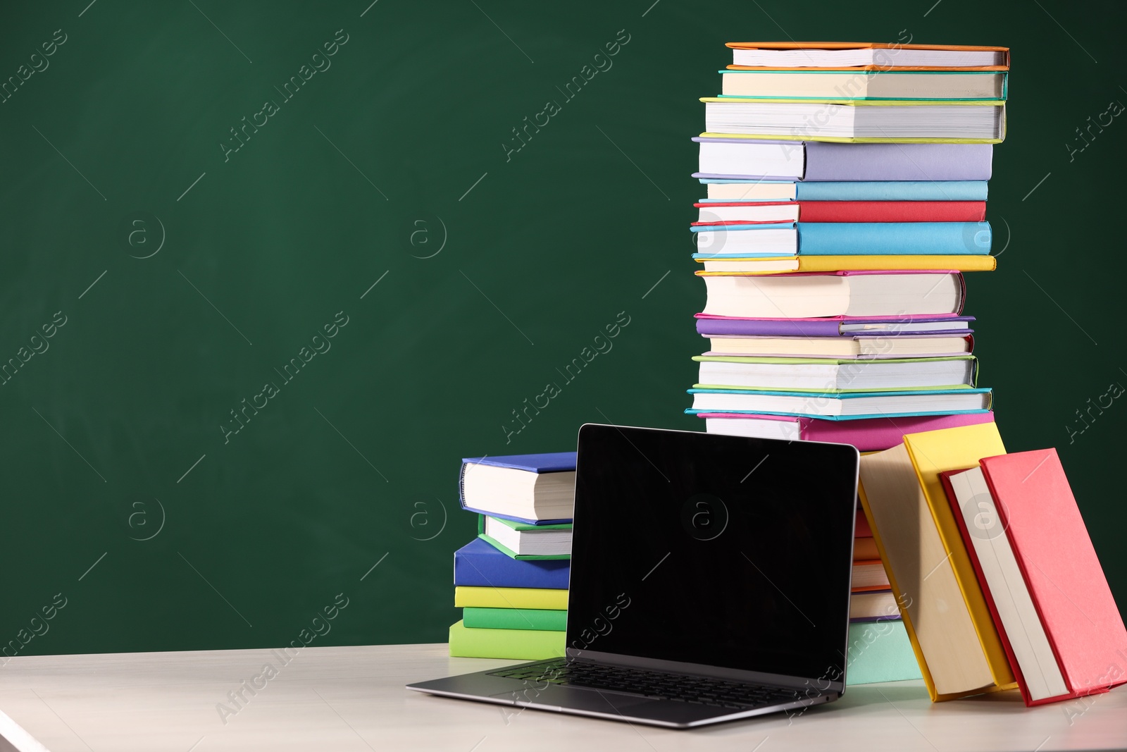 Photo of Stacks of many colorful books and laptop on white table near chalkboard in classroom. Space for text