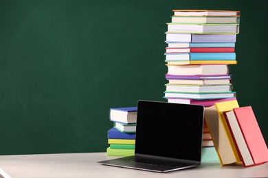 Stacks of many colorful books and laptop on white table near chalkboard in classroom. Space for text