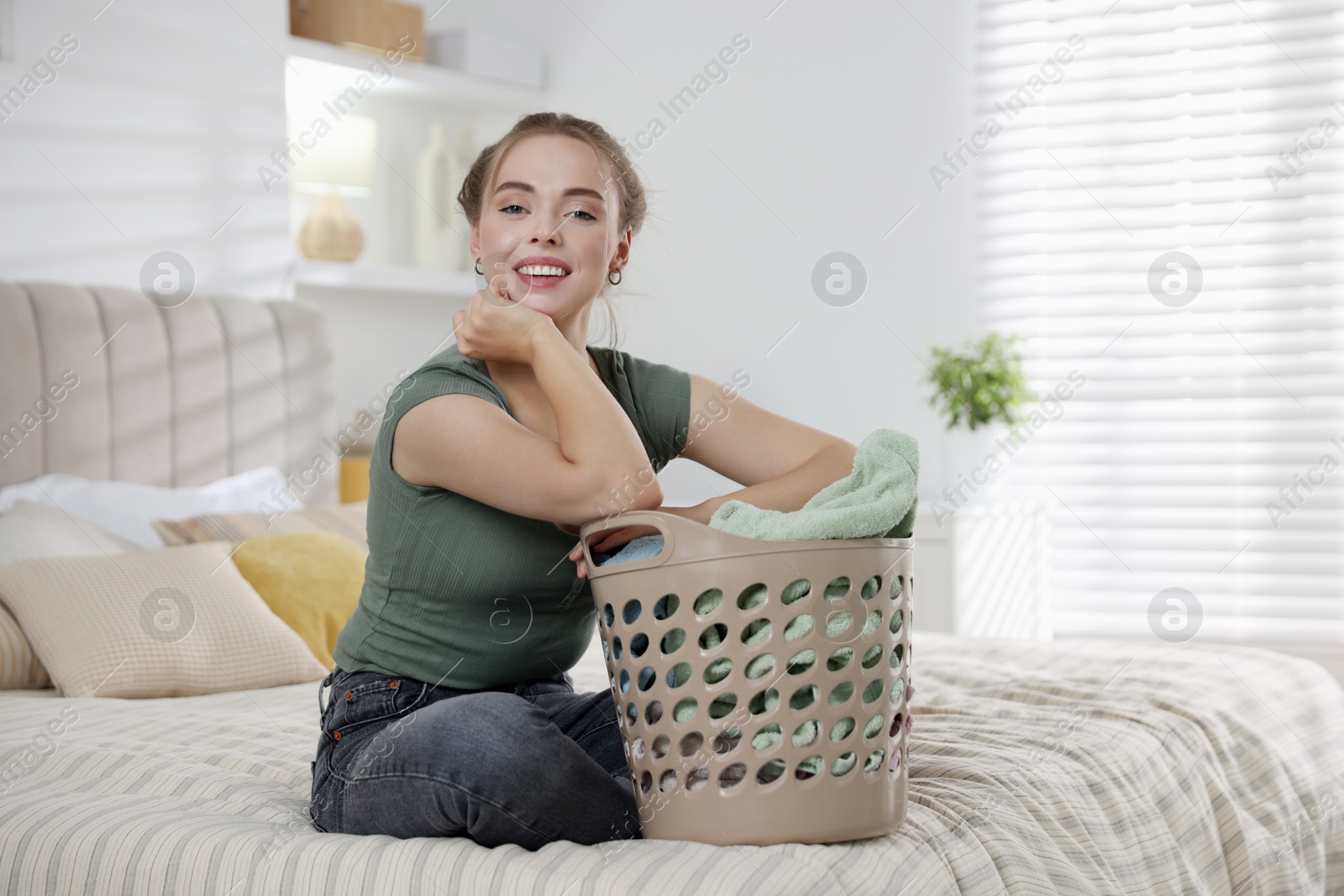 Photo of Happy young housewife with basket full of laundry on bed at home