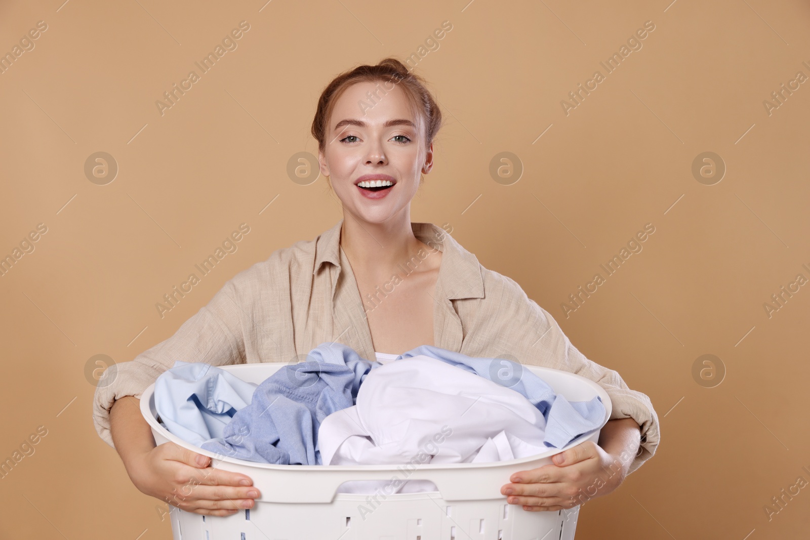 Photo of Happy young housewife with basket full of laundry on pale orange background