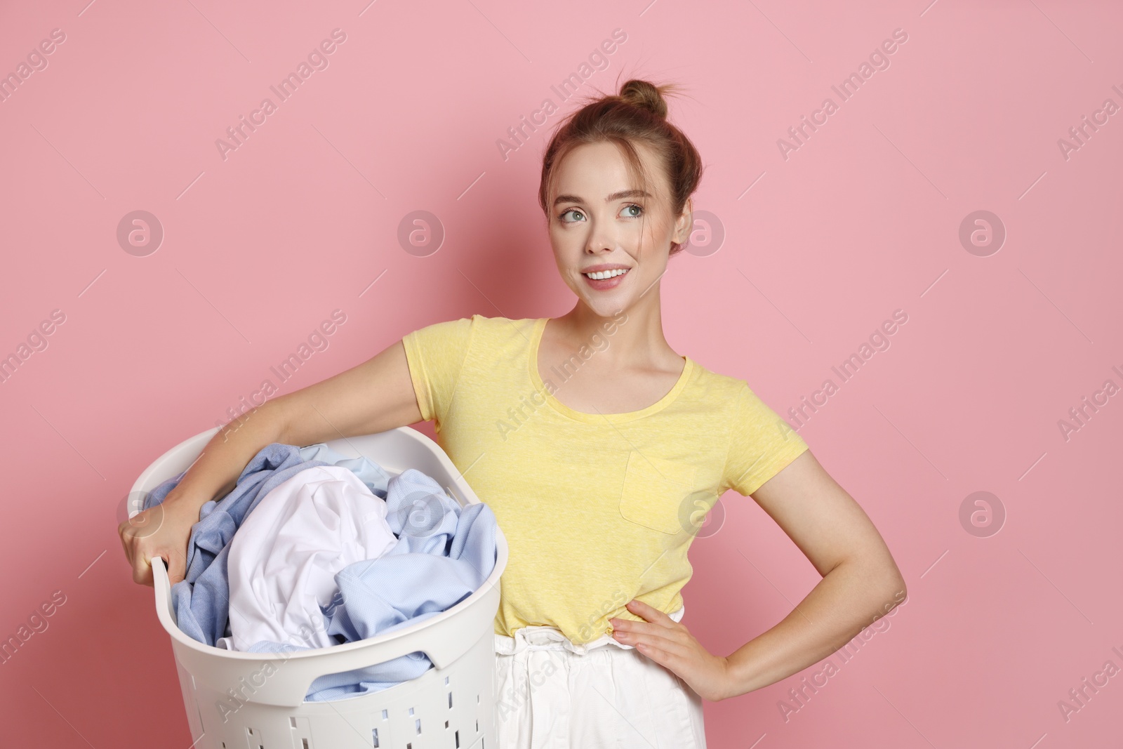 Photo of Happy young housewife with basket full of laundry on pale pink background