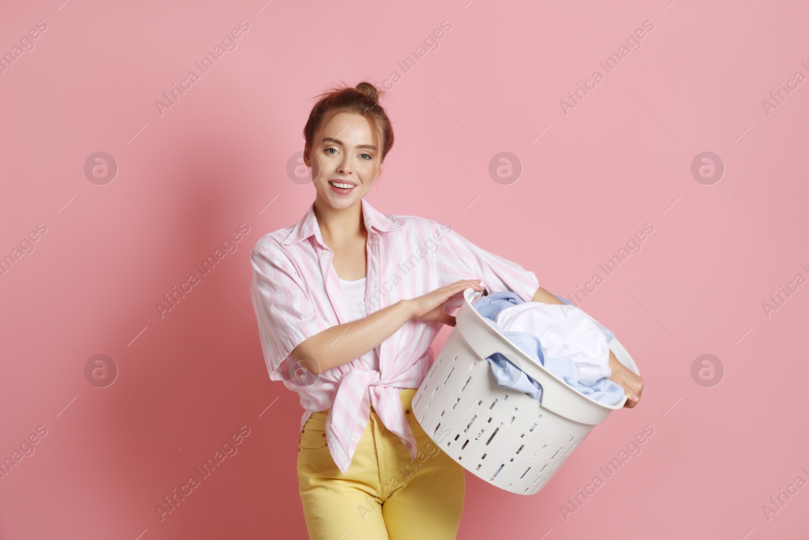 Photo of Happy young housewife with basket full of laundry on pale pink background