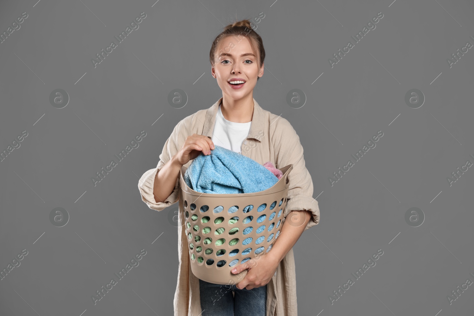 Photo of Happy young housewife with basket full of laundry on grey background