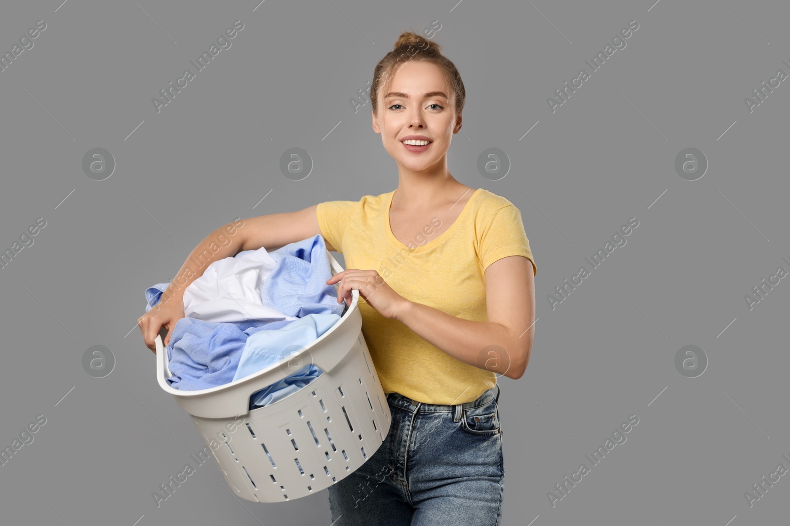 Photo of Happy young housewife with basket full of laundry on grey background. Space for text