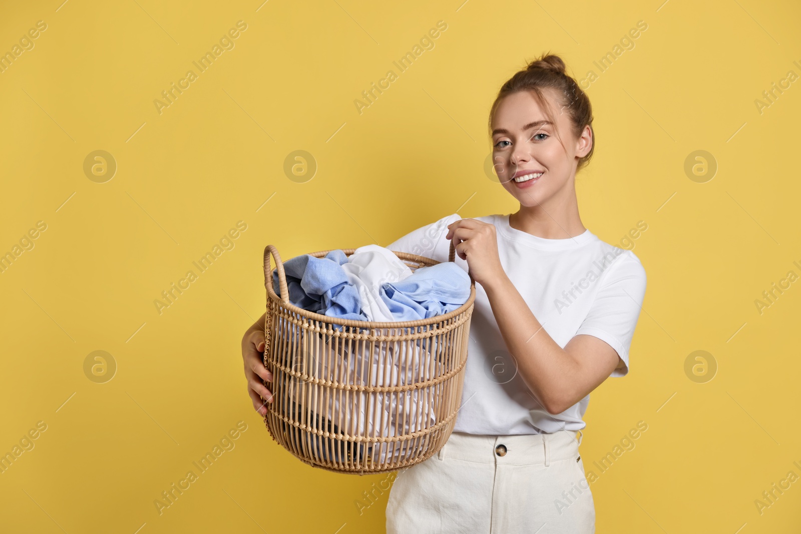 Photo of Happy young housewife with basket full of laundry on yellow background