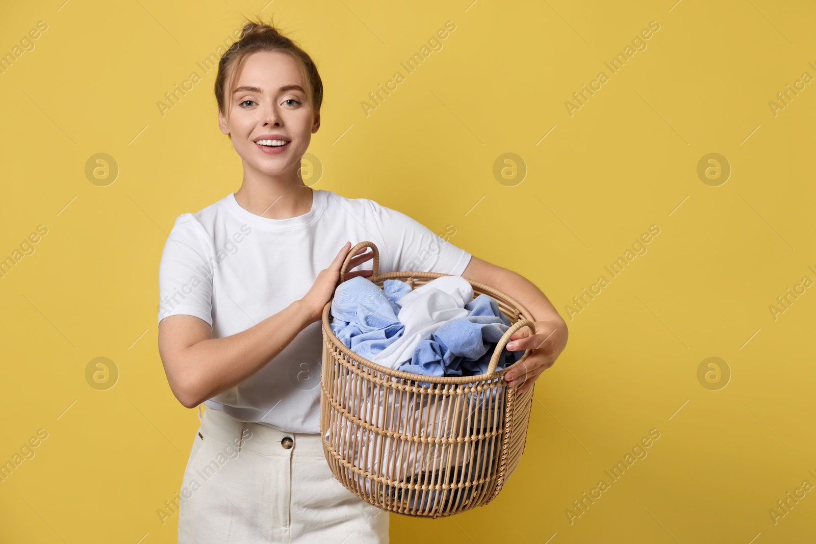 Photo of Happy young housewife with basket full of laundry on yellow background