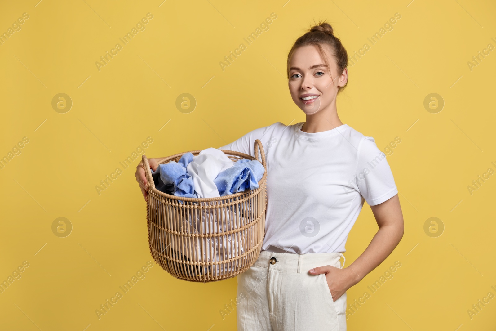 Photo of Happy young housewife with basket full of laundry on yellow background