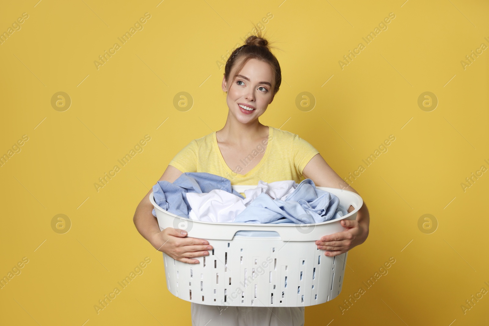 Photo of Happy young housewife with basket full of laundry on yellow background