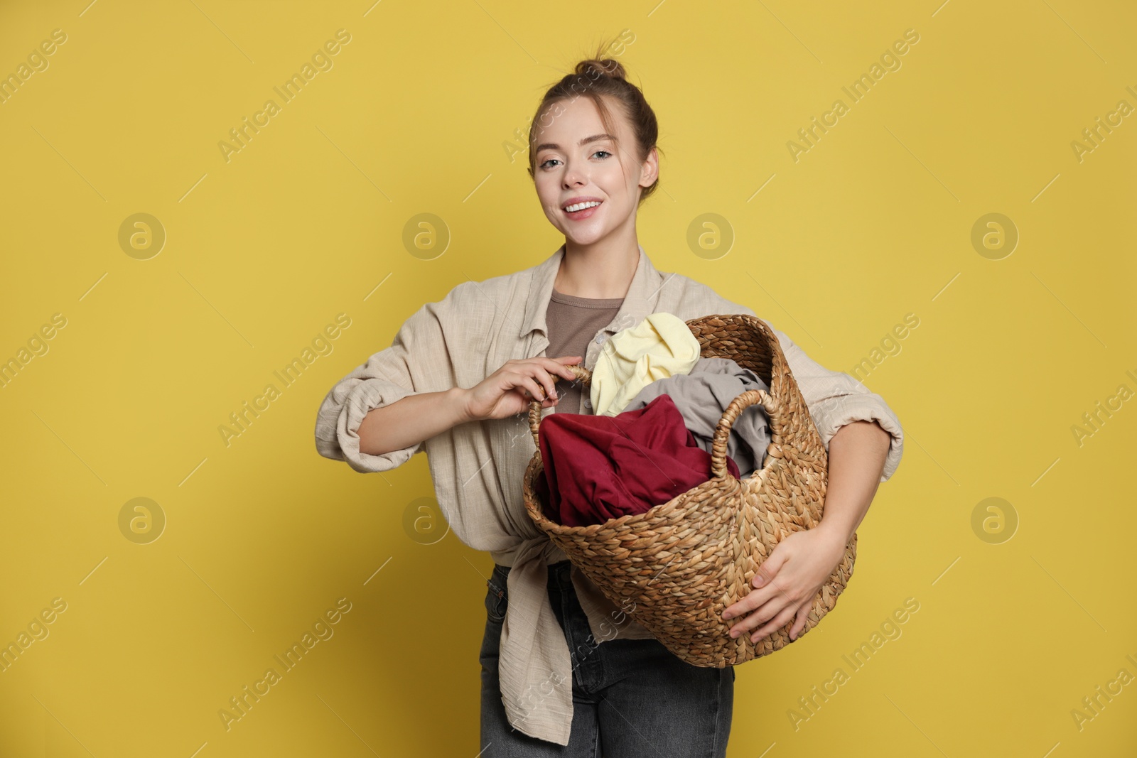 Photo of Happy young housewife with basket full of laundry on yellow background