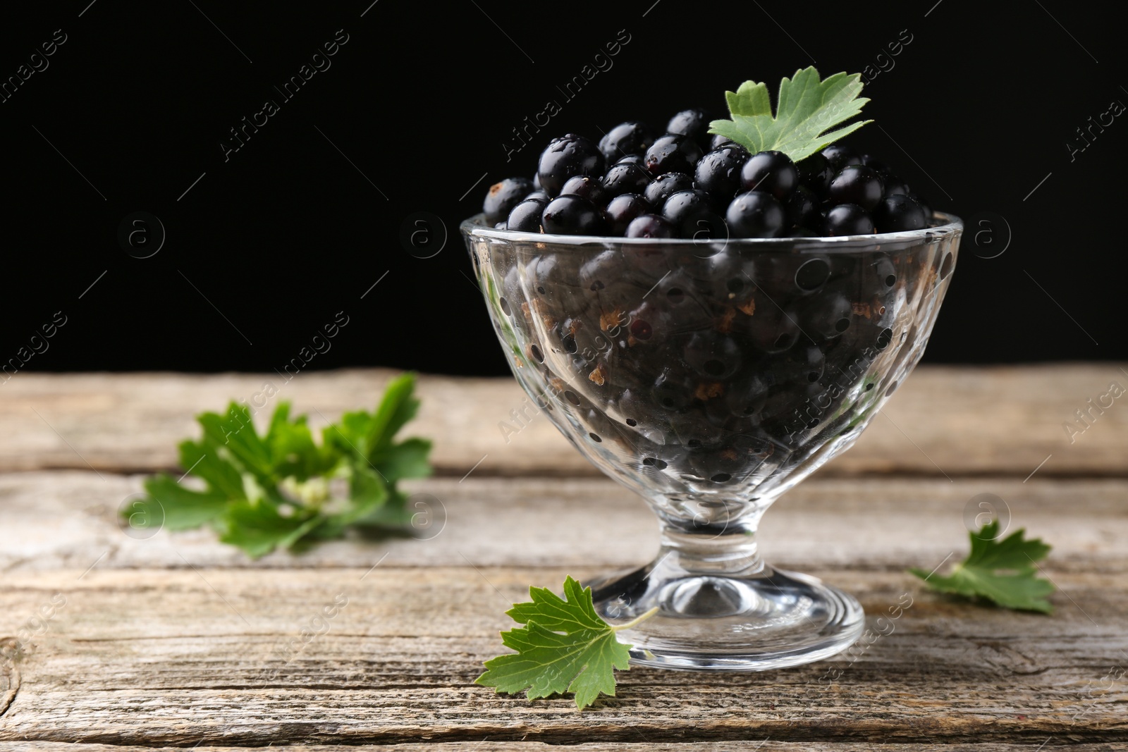 Photo of Ripe black currants and leaves in glass dessert bowl on wooden table, space for text