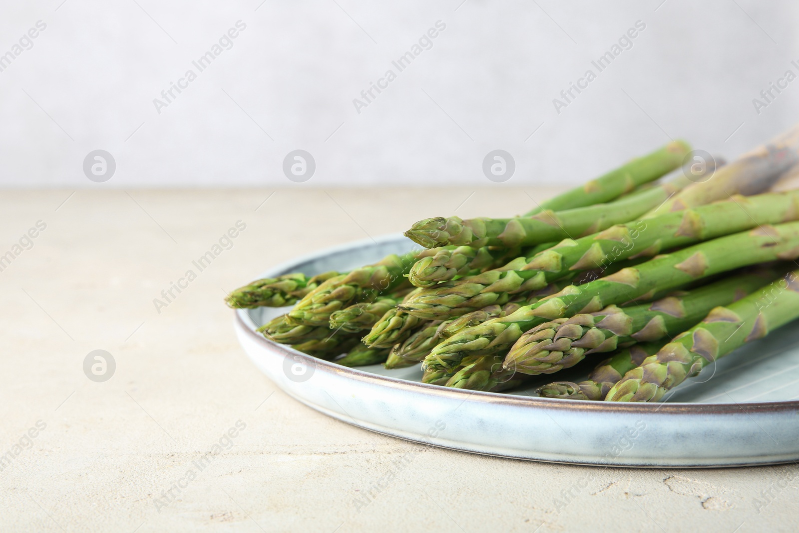 Photo of Plate with fresh green asparagus stems on light textured table, closeup. Space for text