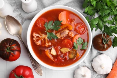 Delicious homemade stew in bowl, spoon and ingredients on white tiled table, flat lay