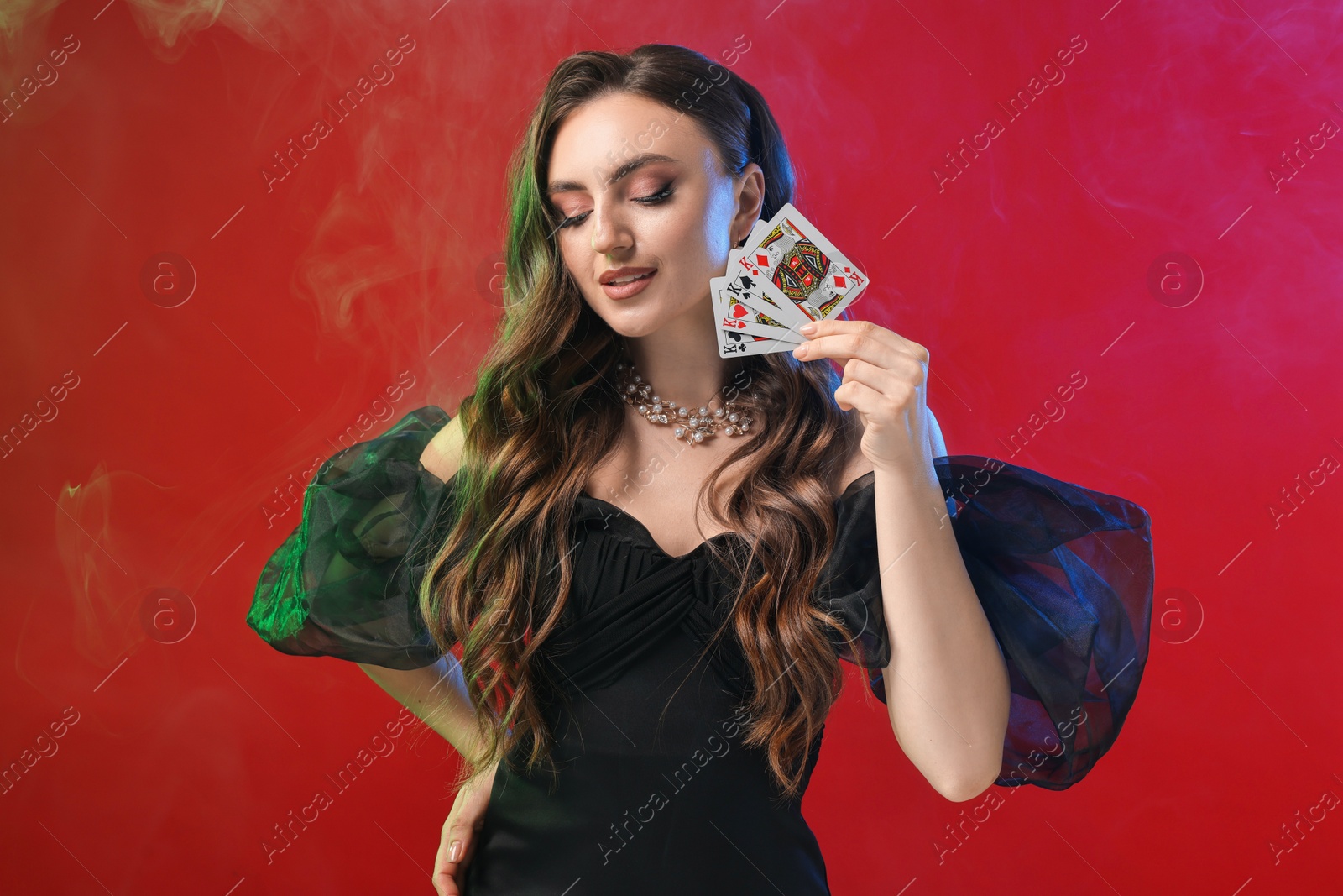 Photo of Poker game. Charming woman with playing cards on red background in color lights and smoke