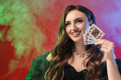 Poker game. Smiling woman with playing cards on red background in color lights and smoke. Space for text