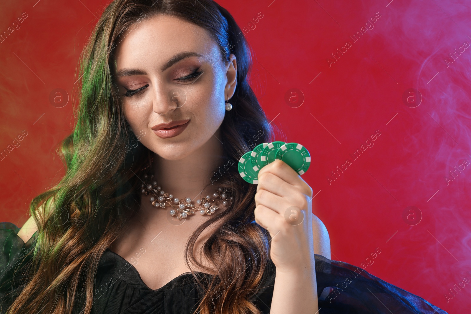 Photo of Charming woman with poker chips on red background in color lights and smoke. Space for text