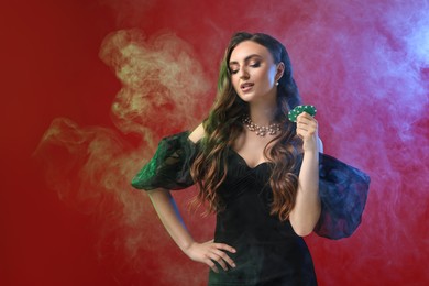 Photo of Charming woman with poker chips on red background in color lights and smoke. Space for text