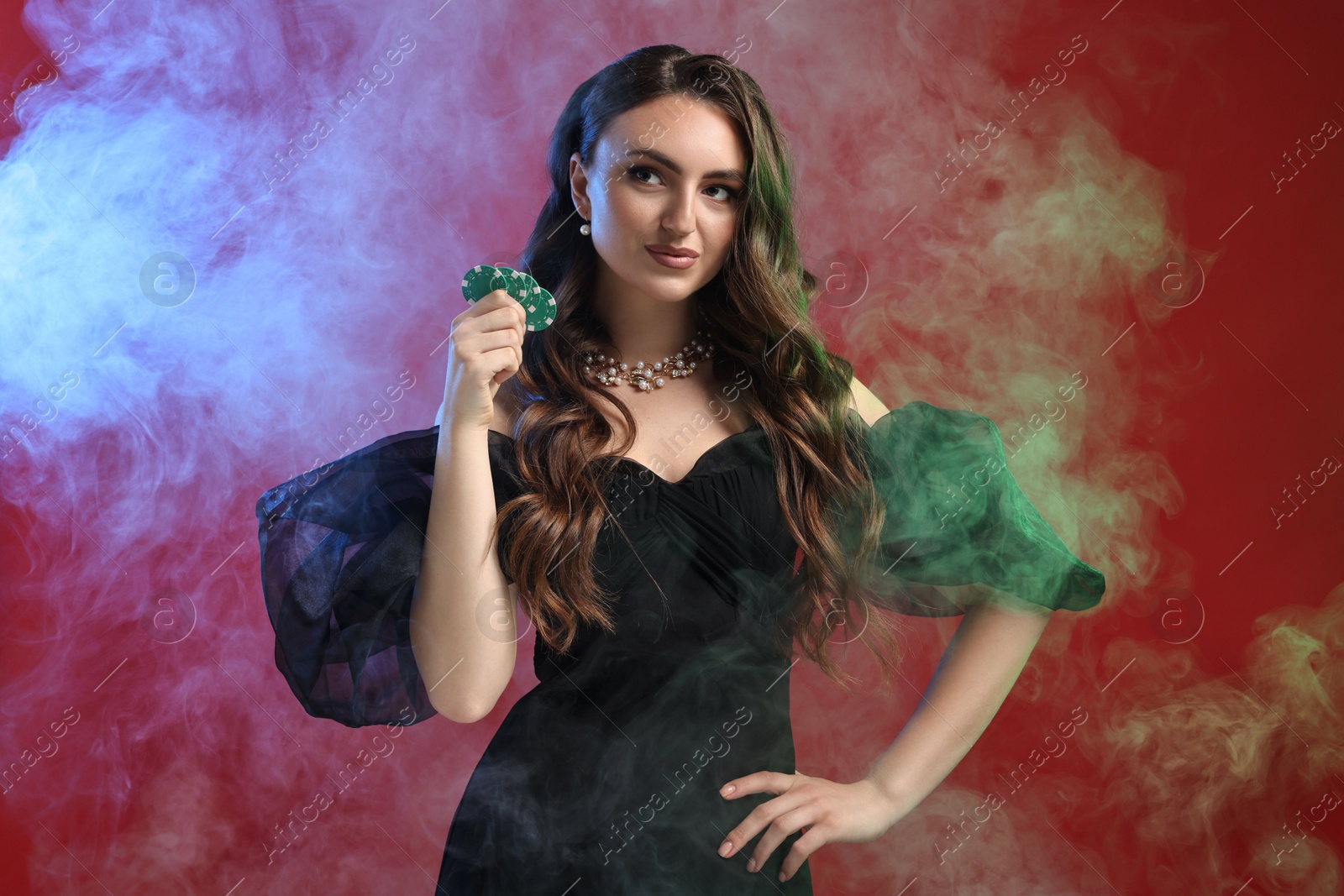 Photo of Charming woman with poker chips on red background in color lights and smoke