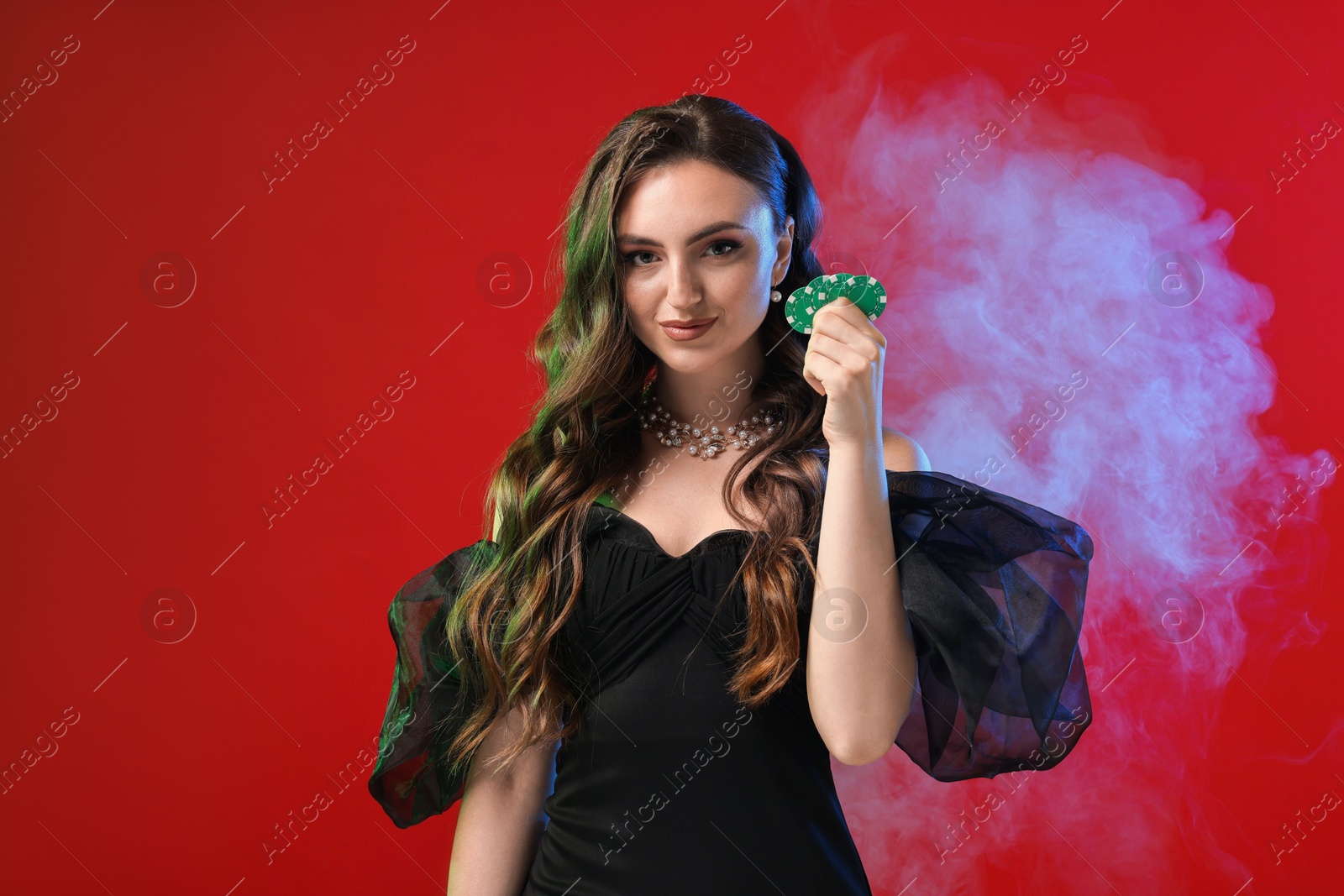 Photo of Charming woman with poker chips on red background in color lights and smoke