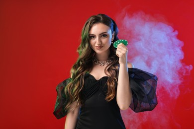 Photo of Charming woman with poker chips on red background in color lights and smoke