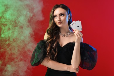 Poker game. Charming woman with playing cards on red background in color lights and smoke