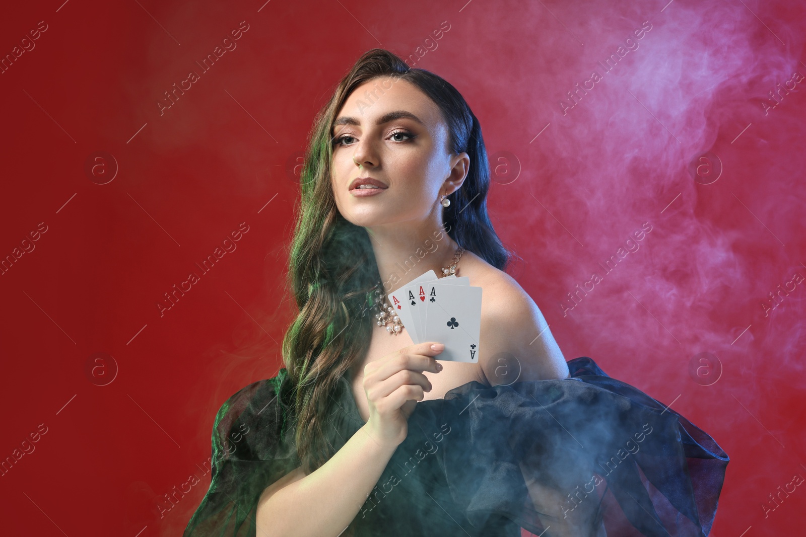 Photo of Poker game. Charming woman with playing cards on red background in color lights and smoke