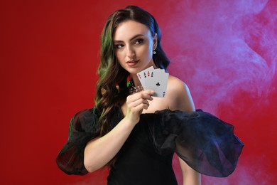 Photo of Poker game. Charming woman with playing cards on red background in color lights and smoke