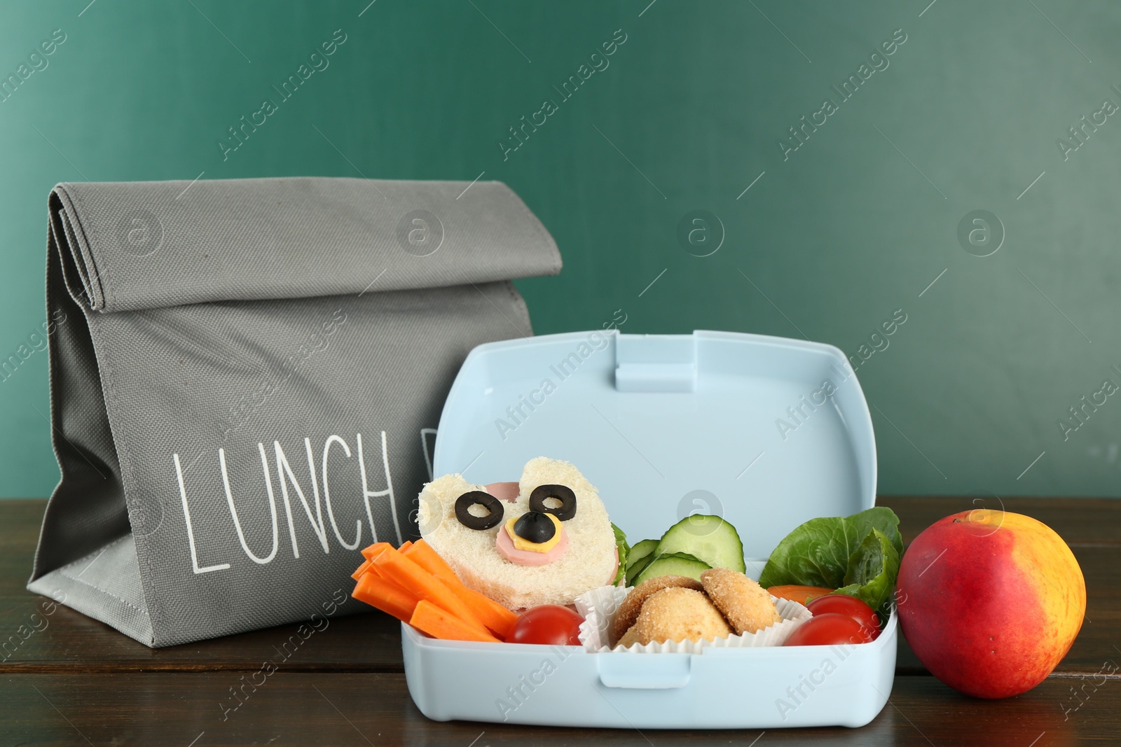 Photo of Gray bag, lunch box with sandwich, fresh vegetables, cookies and peach on wooden table against color background