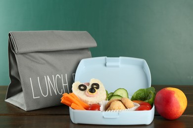 Photo of Gray bag, lunch box with sandwich, fresh vegetables, cookies and peach on wooden table against color background