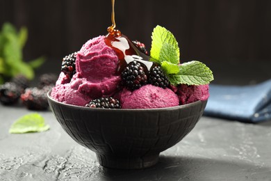 Pouring syrup onto delicious blackberry sorbet in bowl at gray textured table, closeup