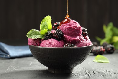 Photo of Pouring syrup onto delicious blackberry sorbet in bowl at gray textured table, closeup