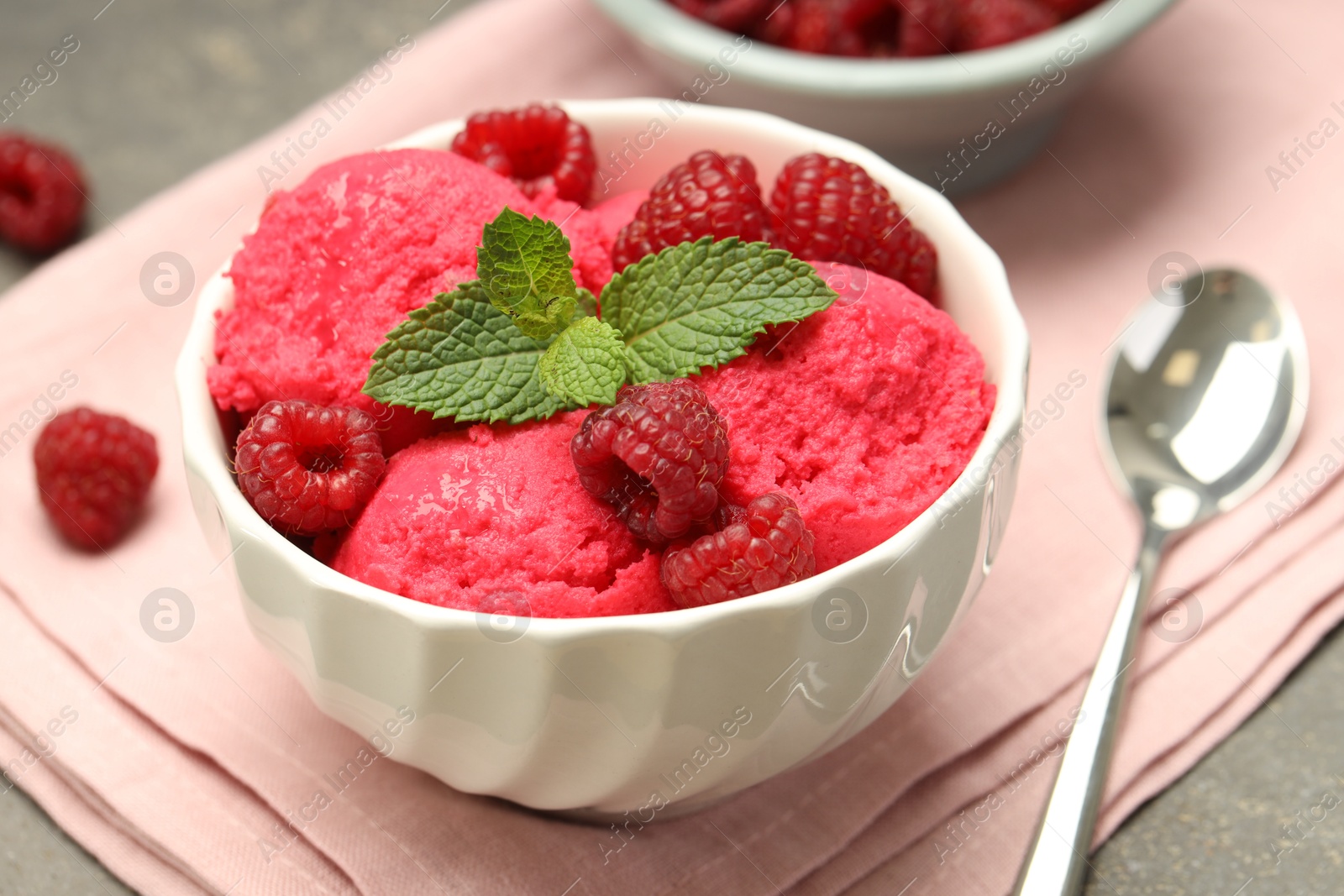 Photo of Delicious raspberry sorbet with fresh berries in bowl and spoon on gray textured table, closeup