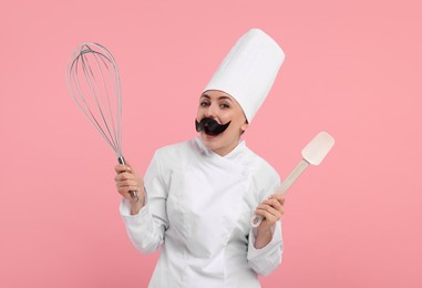 Funny pastry chef with fake mustache holding big whisk and spatula on pink background