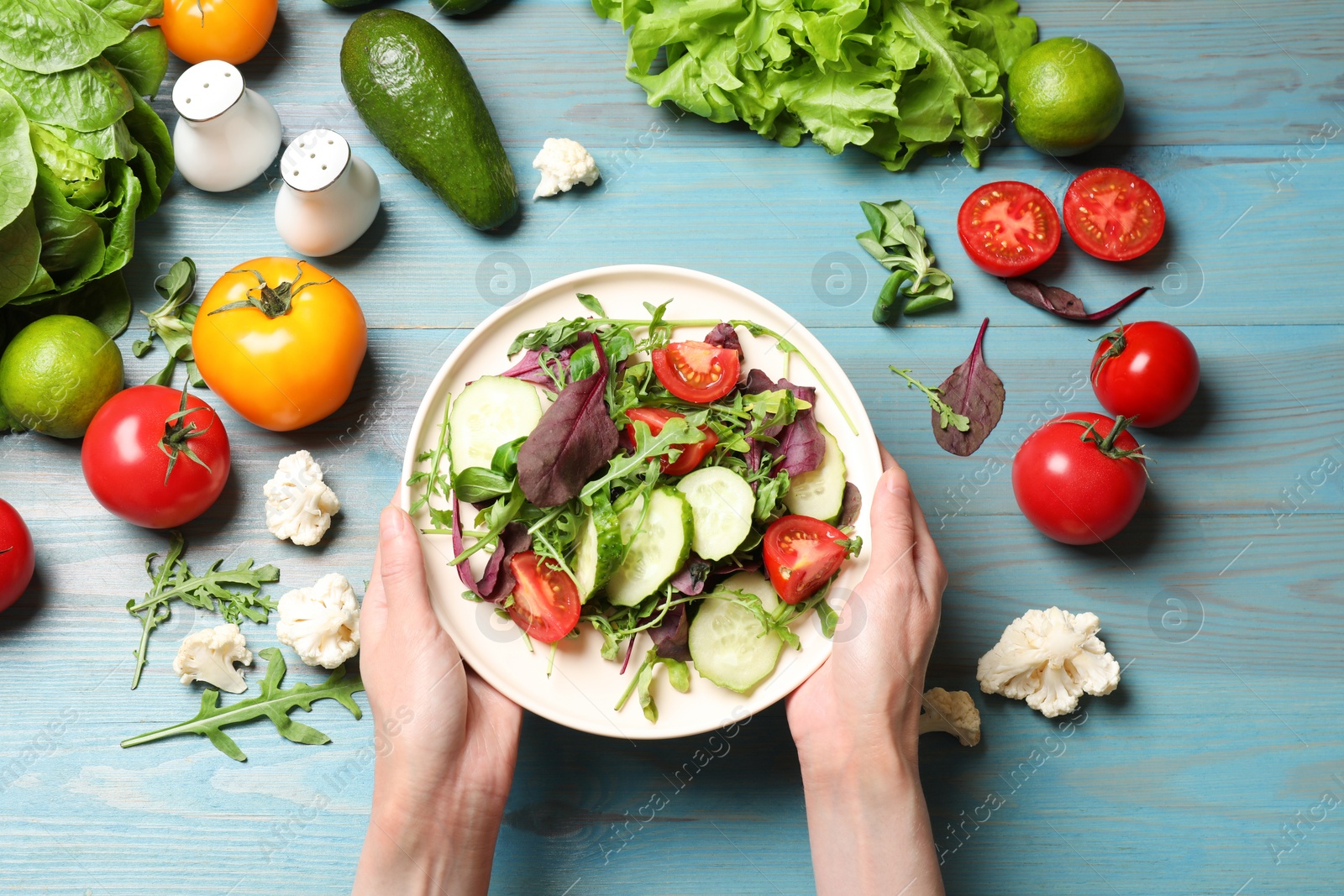 Photo of Healthy vegetarian food. Woman holding plate of salad at light blue wooden table with vegetables, top view