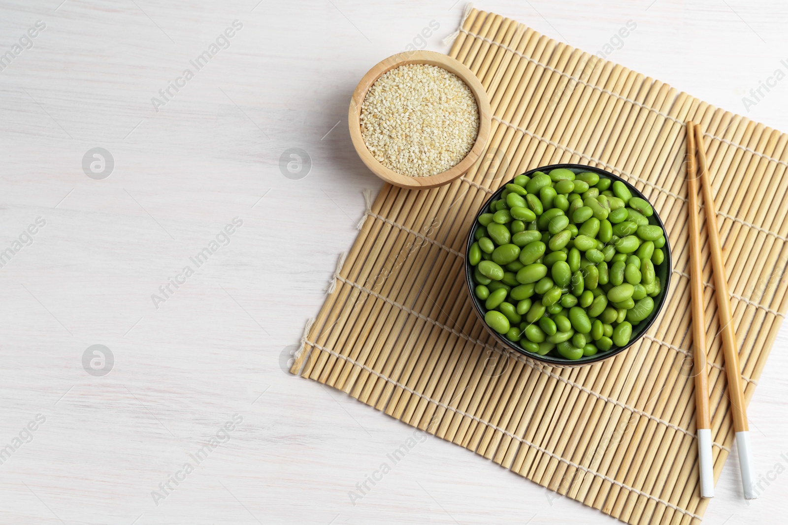 Photo of Fresh edamame soybeans in bowl, chopsticks and sesame seeds on white wooden table, top view. Space for text