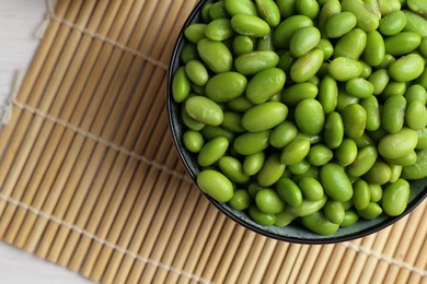 Fresh edamame soybeans in bowl and bamboo mat on light table, top view