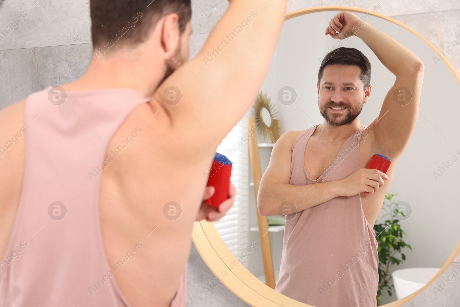 Photo of Smiling man applying solid deodorant near mirror at home, back view