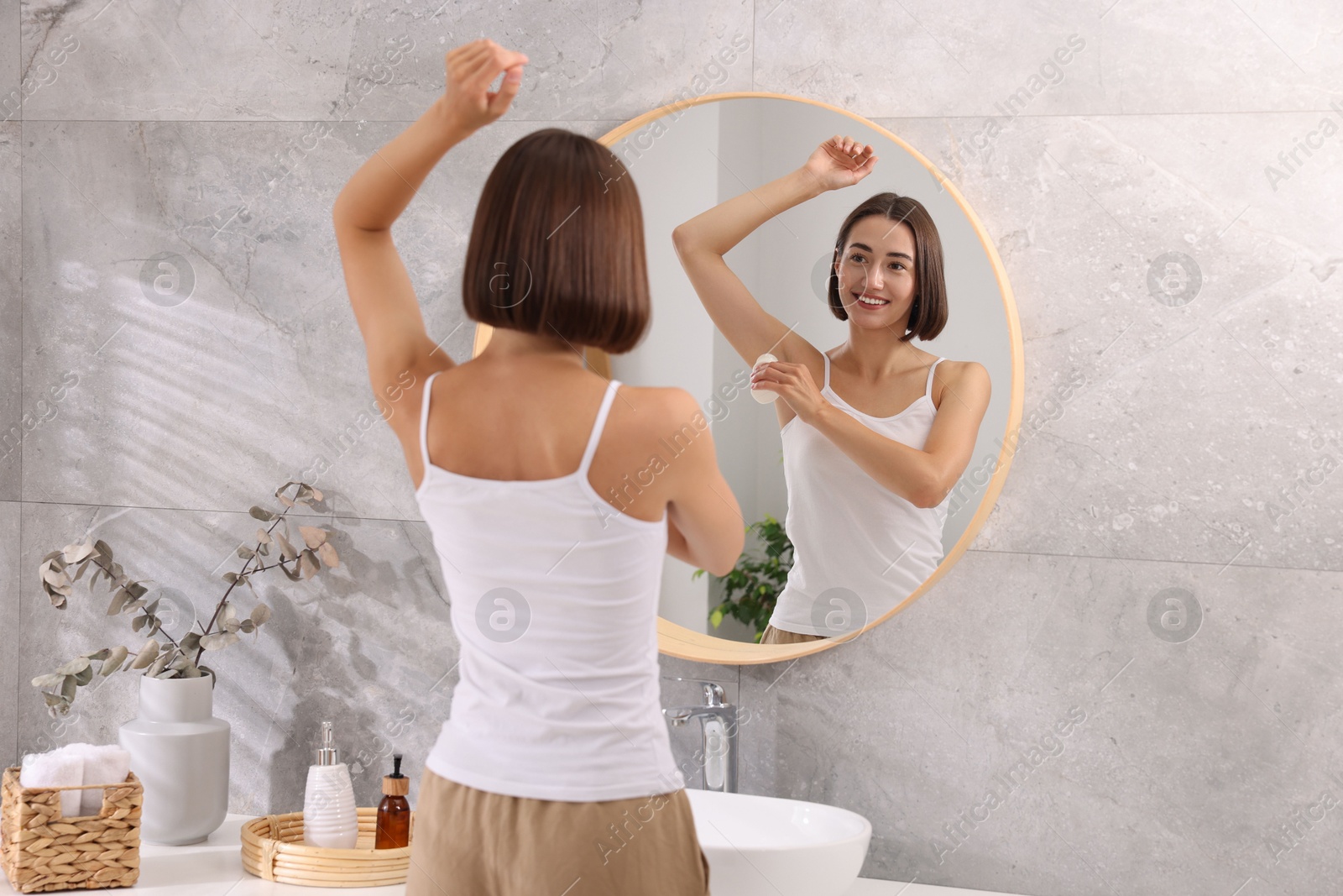 Photo of Smiling woman applying roll-on deodorant near mirror at home, back view