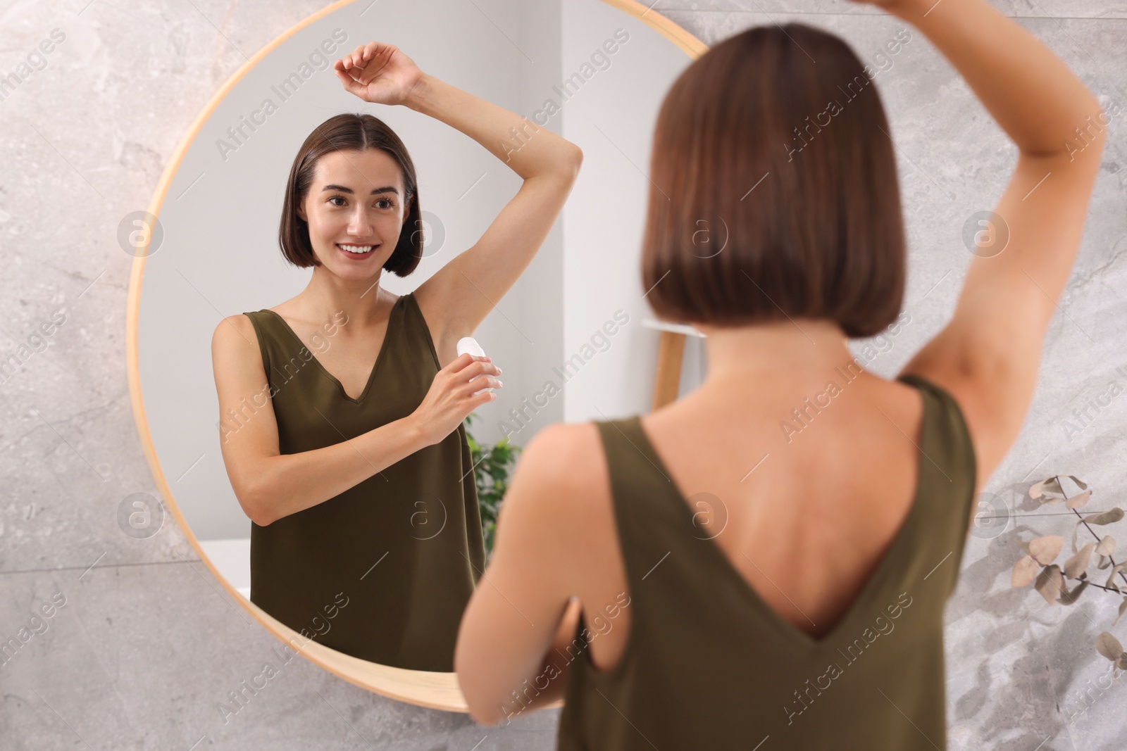 Photo of Smiling woman applying roll-on deodorant near mirror at home, back view