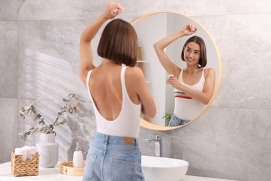 Smiling woman applying roll-on deodorant near mirror at home, back view