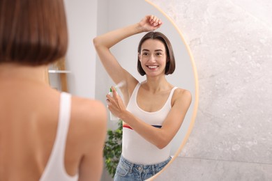 Smiling woman applying spray deodorant near mirror at home