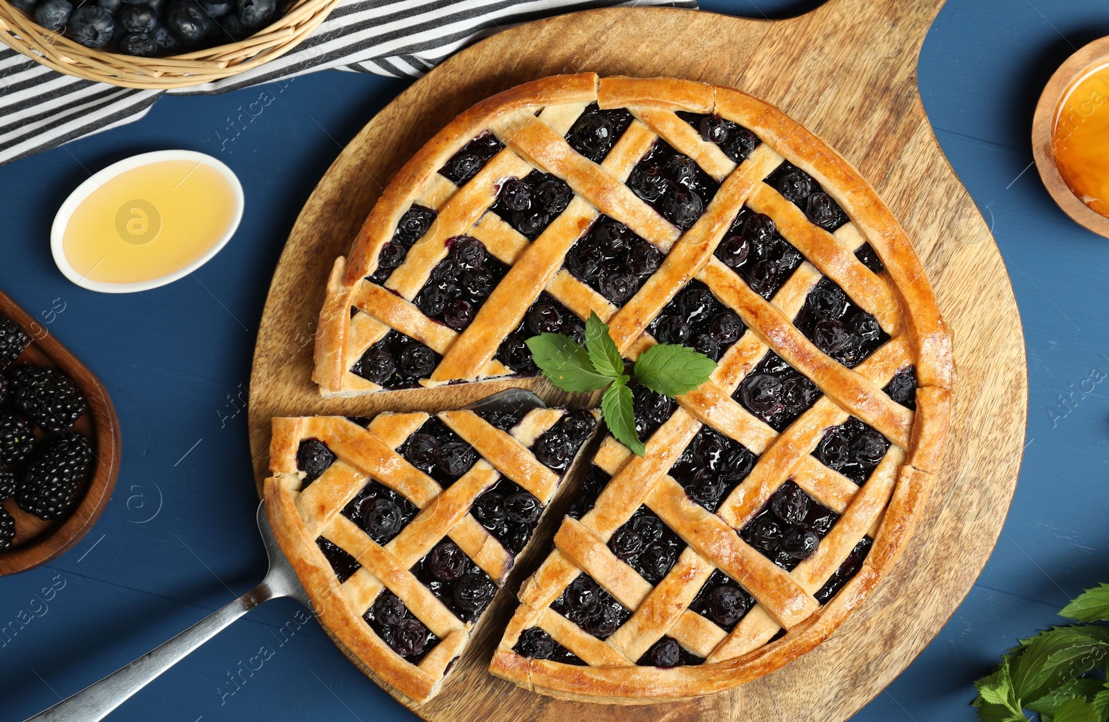 Photo of Tasty homemade pie with blueberries, fresh berries, mint, honey and cake server on blue wooden table, flat lay