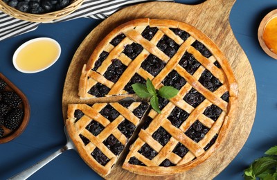 Photo of Tasty homemade pie with blueberries, fresh berries, mint, honey and cake server on blue wooden table, flat lay