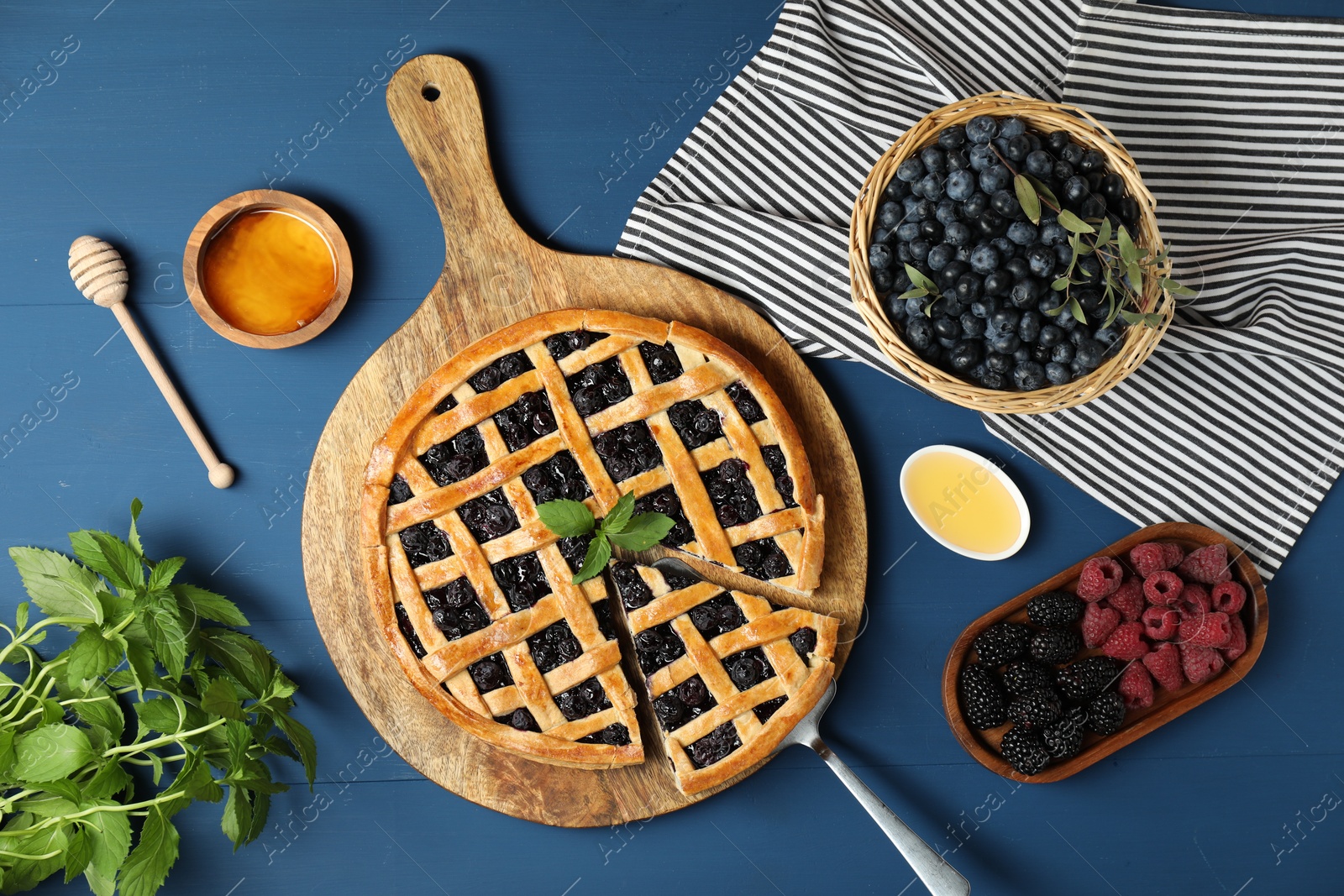 Photo of Tasty homemade pie with blueberries, fresh berries, mint, honey and cake server on blue wooden table, flat lay