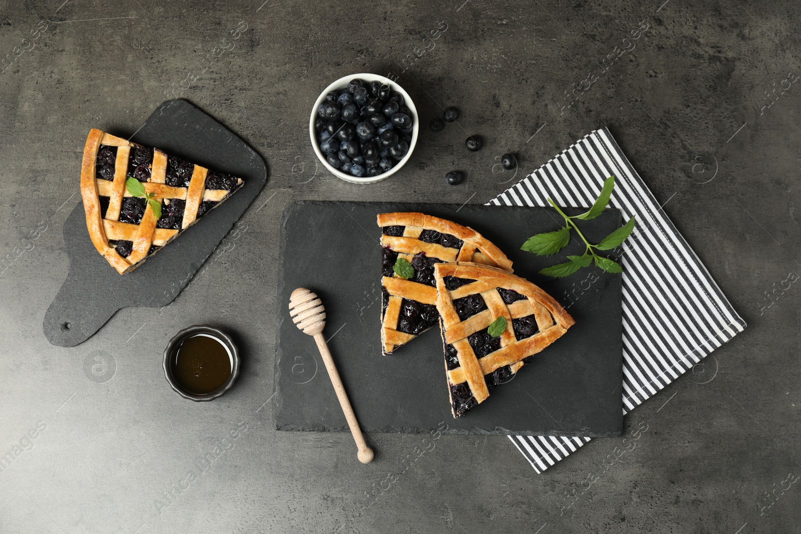 Photo of Pieces of tasty homemade pie with blueberries, mint and honey on gray textured table, flat lay