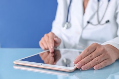 Doctor with tablet at table against blue background, closeup view