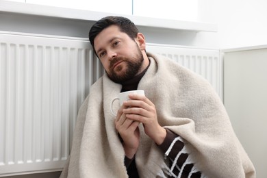 Man with cup of hot drink near heating radiator at home