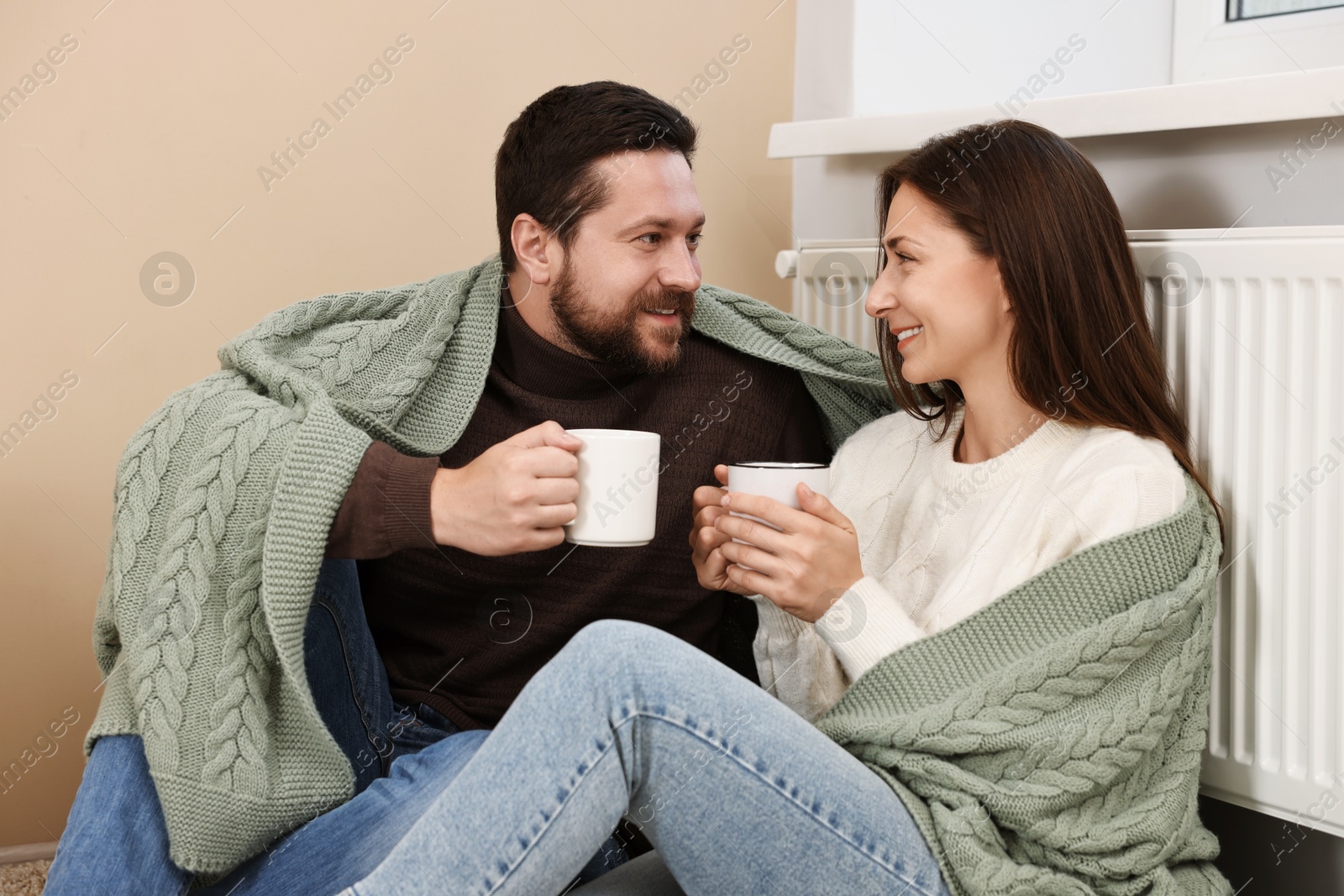 Photo of Happy couple with cups of hot drinks near heating radiator at home