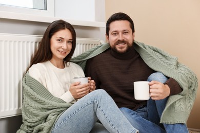 Photo of Happy couple with cups of hot drinks near heating radiator at home